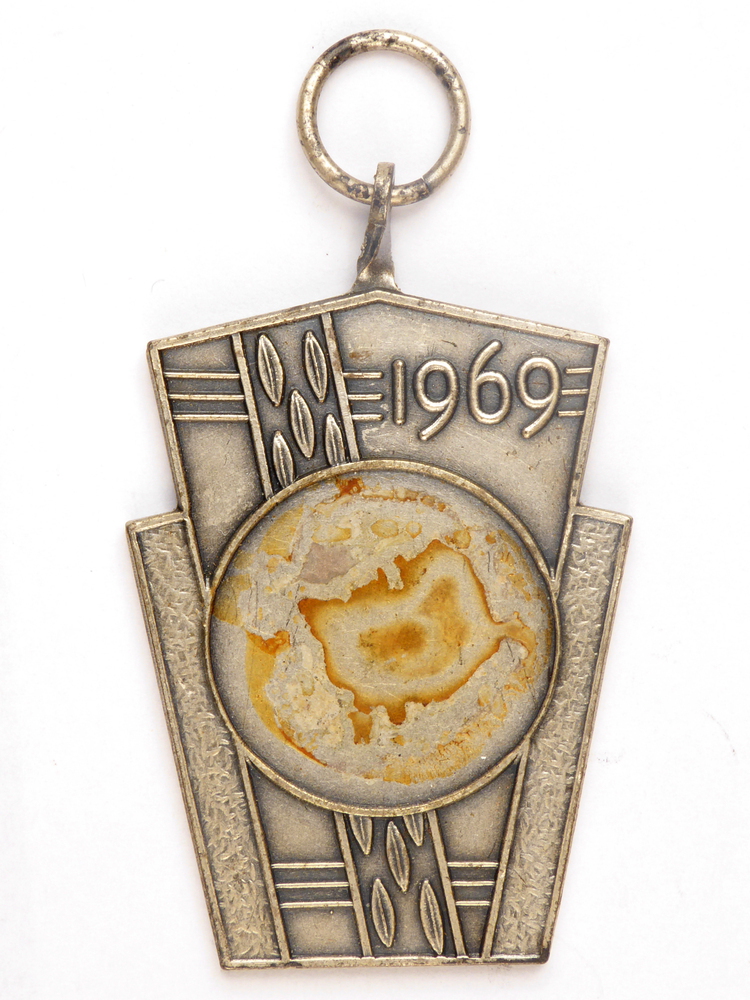 zilverkleurige medaille met op voorkant afbeelding bladeren, middenstuk met afbeelding geit ontbreekt, op achterkant inscriptie VW